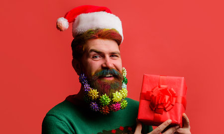 Smiling bearded man in Santa hat with gift box. New Year present. Christmas decorated beardの写真素材