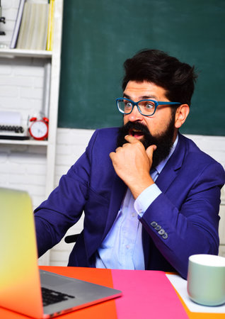 Surprised businessman in suit sitting in modern office looking at laptop happy by good news. Shocked bearded man in glasses celebrating project success. Male teacher or student at desk in classroom.の写真素材