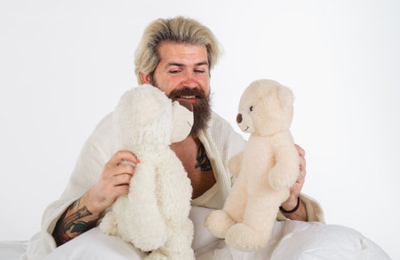 Man with two teddy bears. Bearded man playing in bed with teddy bears. Smiling hipster in bathrobe with cute teddy bear toy. Soft toy for sleeping. Handsome guy in pajamas in bedroom with teddy bears.の写真素材