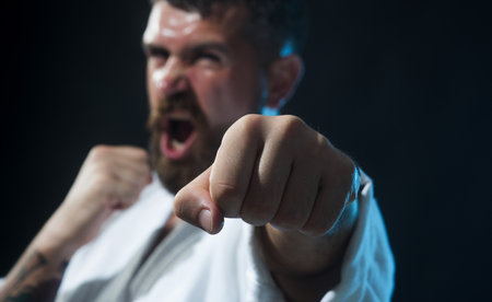 Karate man in fighting stance. Screaming karate fighter practicing punches in gym, selective focus on fist. Angry bearded man in kimono with hand clenched in fist. Karate man exercising combat sport.の写真素材