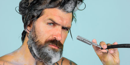 Bearded man shaving beard with straight razor in shower. Beard care. Closeup portrait of shaving man. Bearded man with vintage razor for shaving. Classic shave. Barbershop advertising. Banner design.の写真素材