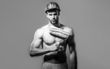 Painter man with painting roller. Strong male painter, designer or decorator with naked torso holds paint roller. Serious shirtless builder in protective hard hat with painting tools. Black and whiteの写真素材