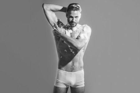 Muscular sexy man in boxer shorts taking shower in bathroom. Handsome man washing body with cleansing foam and sponge. Black and white. Attractive guy takes shower with moisturizing gel. Body careの写真素材