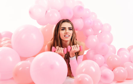 Fitness, sport, healthy lifestyle. Sporty woman with dumbbells lying between pink balloons. Sensual girl with long hair and perfect makeup with dumbbells. Personal training. Weight training. Workoutの写真素材