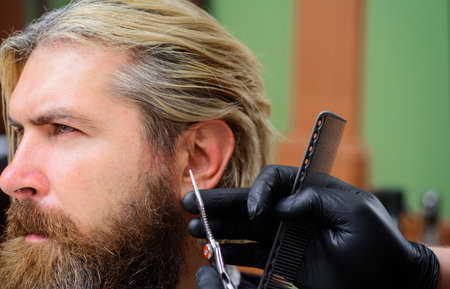 Hairdresser works with client in barbershop. Hairdresser, hair salon. Men's hairstylist. Barber comb, scissors. Work in barber shop. Bearded man visits hairdresser in barbershop. Beard care. Haircutの写真素材