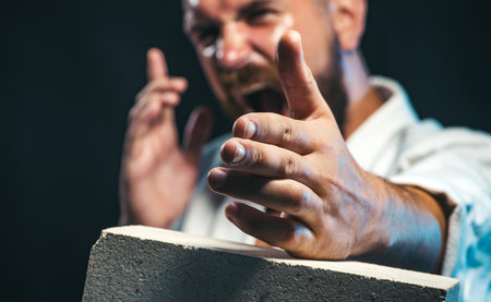 Karate man in kimono shows kick by hand breaking concrete slab. Karate master instructor breaks brick, selective focus on hand. Man hand breaking concrete brick. Karate fighter smashes concrete slabの写真素材