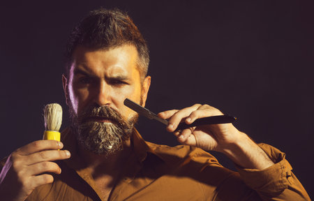 Barbershop accessories. Bearded man with straight razor and shaving brush. Beard care. Barber with vintage razor for shaving. Handsome guy with beard in barbershop. Hairdresser with tools for shavingの写真素材