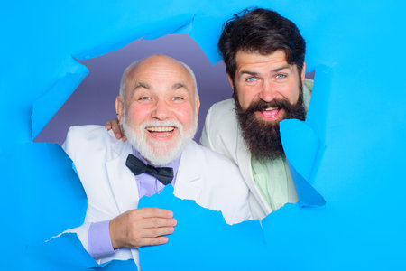 Successful bearded men looking through hole in paper. Happy adult son and senior old father in white suits. Happy Father's day. Family relationship. Fashionable clothes.の写真素材