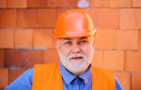 Construction worker in safety helmet near brick wall on construction site. Male builder in protective hard hat working at object. Portrait of architect or civil engineer in work clothes at workplaceの写真素材