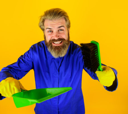 Professional cleaning service. Cleaner man with sweep brush and dustpan. Cleaning agency. Smiling bearded man in uniform with dustpan and whisk broom. Cleaning equipment, sweeping tools. Cleaning dayの写真素材