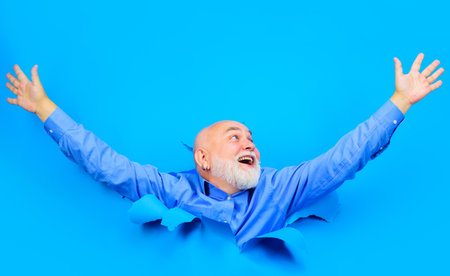 Happy old bearded man looking through hole in paper. Smiling senior man in stylish shirt raised hands. Handsome male peeking from ripped paper hole. Season sales, discount. Copy space for advertising.の写真素材