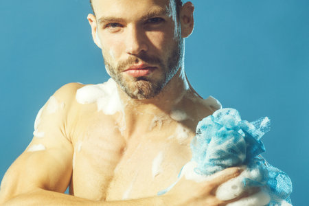 Muscular sexy man taking shower in bathroom. Closeup portrait handsome young man washing body with cleansing foam and sponge. Body care and skincare. Attractive guy takes shower with moisturizing gel.の写真素材