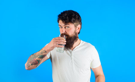 Handsome bearded man drinking glass of fresh water. Attractive man in casual white t-shirt drinking filtered water in morning. Healthy lifestyle, good life habits, water balance, weight loss and diet.の写真素材