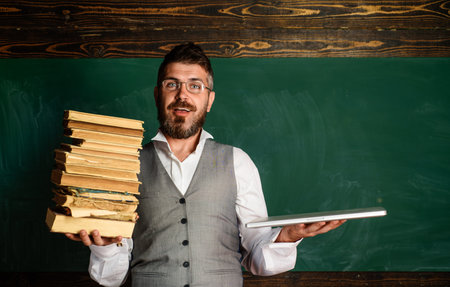 Teacher in glasses with laptop and textbook in classroom. Smiling student with laptop and books in front blackboard. E-learning, e-book and e-library. Choice between paper books and e-learning gadgetの写真素材
