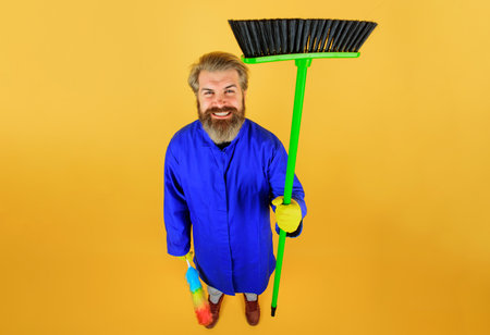 Cleaner man with broom. Professional cleaning service. Full length portrait of cleaner with sweeping tools and equipment. Cleaning agency. Smiling bearded man in uniform and rubber gloves with broom.の写真素材