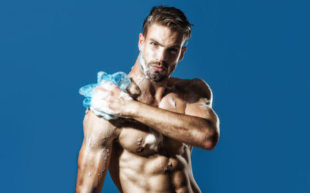 Muscular sexy man taking shower in bathroom. Attractive serious guy takes shower with moisturizing gel. Body care, skincare and spa. Handsome athlete man washing body with cleansing foam and sponge.の写真素材