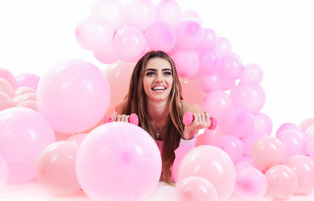 Fitness, sport and healthy lifestyle. Sporty woman with dumbbells lying between pink balloons. Smiling young girl with long hair and perfect makeup with dumbbells. Personal training. Weight trainingの写真素材