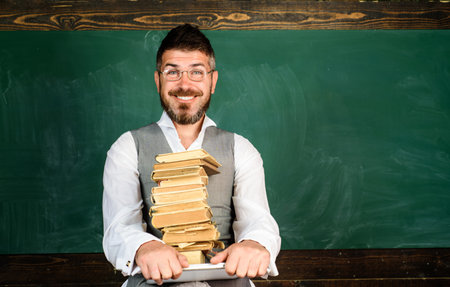 Teacher in glasses with laptop and pile textbook in classroom. Smiling student with stack of books on laptop in front blackboard. Bearded teacher giving lesson to students. Copy space for advertisingの写真素材