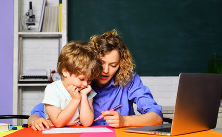 Female elementary school teacher helping cute schoolboy with homework. Knowledge, education and learning. Primary school pupil and private tutor working together at table in classroom. Back to schoolの写真素材