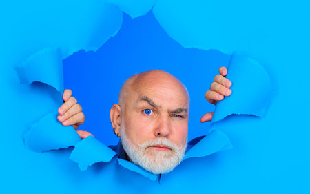 Serious old bearded man looking through hole in paper. Handsome male breaking through paper wall. Senior man with blue lenses peeping through hole in paper. Discount, sale, season sales. Black Fridayの写真素材