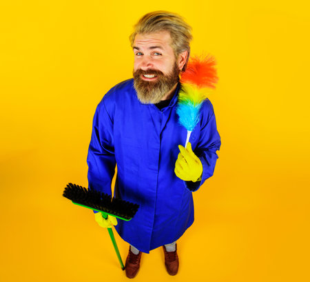 Smiling bearded man in uniform with broom and dust brush full length portrait. Professional cleaning service. Cleaner in rubber gloves with cleaning supplies. Male worker with broom and pp-duster.の写真素材