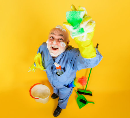 Big cleaning time. Housecleaning. Full length portrait of smiling old man in uniform with cleaning equipment. Professional cleaning service. Cleaner in rubber gloves with sponge and cleaning suppliesの写真素材