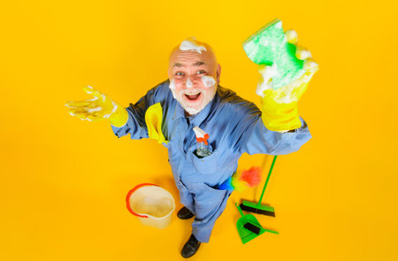 Big cleaning time. Full length portrait of smiling old man in uniform with cleaning equipment. Professional cleaning service. Cleaner in rubber gloves with sponge and cleaning supplies. Housecleaningの写真素材