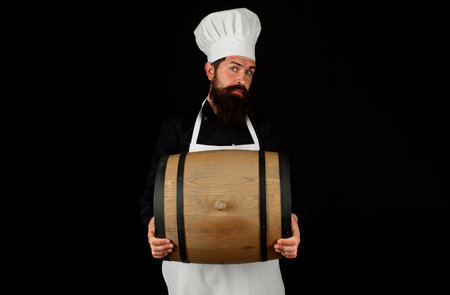 Bearded man with beer barrel. Brewer in apron holds barrel with craft beer. Serious man carrying wooden barrel with wine. Winemaking. Handsome male chef with oak barrel or cask of beer for pub or barの写真素材