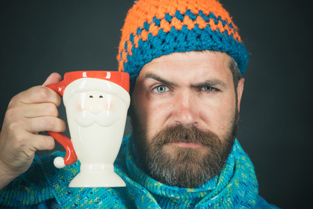 Closeup portrait of serious bearded man in knitted hat and scarf with mug in the shape of Santa. Stylish handsome male with beard and mustache holding mug of hot coffee, cappuccino or teaの写真素材