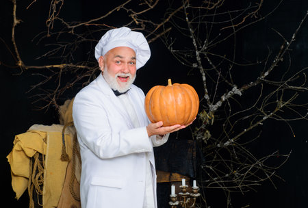 Halloween holiday. Smiling bearded man in chef hat with pumpkin for Halloween. Farm market. Autumn vegetables. Male chef, cook or baker in uniform with pumpkin. Thanksgiving day cooking. Autumn crop.の写真素材