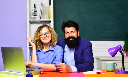 Smiling male and female teachers sitting at desk in classroom. Couple of students preparing for test or exam. Teachers man and woman at college lesson. Student girl with bearded teacher in university.の写真素材