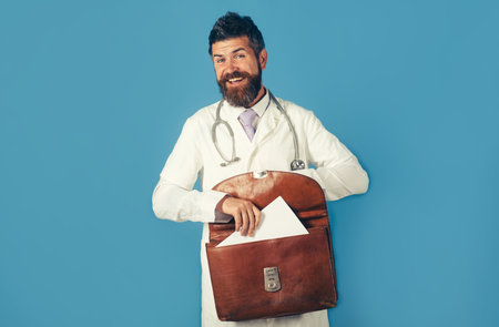 Portrait of smiling male doctor with briefcase. Medical staff. Male doctor holds briefcase and putting some documents inside. Bearded man in medical uniform with stethoscope on neck holds briefcaseの写真素材