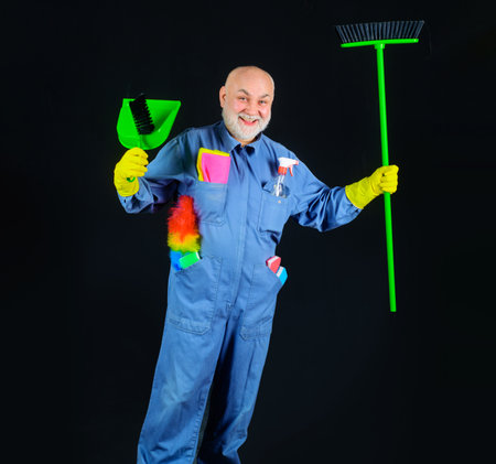Cleaning time. Smiling cleaner with sweeping tools and cleaning supplies. Cleaning agency. Senior man in uniform with broom, dustpan and brush for carpet. Professional cleaning service. Housekeepingの写真素材