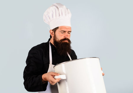 Bearded chef in uniform with large saucepan or pot smelling aroma of freshly cooked food. Cooking food. Man in apron and chef hat holds casserole or cooking pan and sniff aroma dish with eyes closedの写真素材