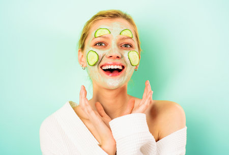 Cosmetology and skincare. Smiling young woman with clay cosmetic face mask and fresh cucumber slices on face. Spa salon, beauty treatment, cosmetic procedures. Natural ingredients. Cucumber face maskの写真素材