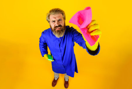 Cleaning service. Male cleaner in rubber gloves with rag and sponge. Full length portrait of bearded man in uniform with cleaning equipment. Housekeeper with pink cloth. Housework and housekeepingの写真素材