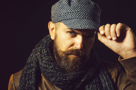 Fashion man. Serious bearded man in leather jacket, knitted scarf and cap. Autumn-winter fashion for men. Bearded hipster in gray scarf adjusting flat cap. Handsome male model in demi-season clothes.の写真素材