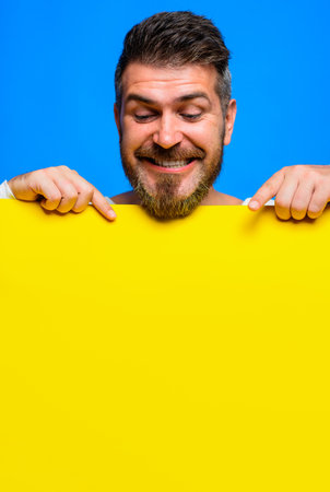 Smiling bearded man pointing with fingers down at advertising board with empty space for text, mockup. Advertising placard, poster. Handsome male with blank signboard, advertising banner.の写真素材