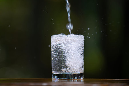 Glass and water with pouring on the green background. Water pouring into glass on wooden table outdoors. Pouring water on nature background. Pouring soda water into glass, closeup.の写真素材