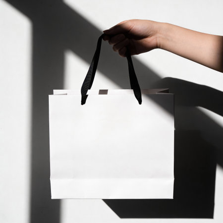 Woman hand holding white paper shopping bag with shadow on white wall background.の写真素材