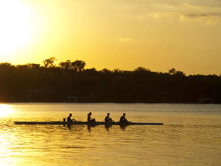 A team of young mean in a row boat silouhetted against setting sunの写真素材