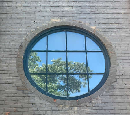 An old round window in a brick wall with the reflection of a tree and blue sky in its pane of glassの写真素材