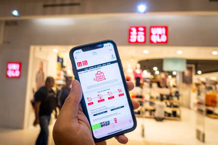 A man's hand holding an iphone X with uniqlo apps infront of Uniqlo store. Uniqlo apps is perfect for those who prefer online shoppingのeditorial素材