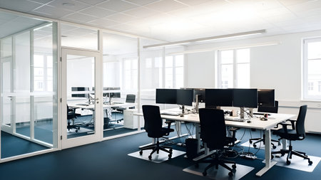 The image depicts a well-organized, modern office space featuring multiple workstations. Each workstation is equipped with a computer monitor, keyboard, and office chair. The office has a clean, minimalist design with white walls, large windows allowing natural light, and a blue carpet. The space appears to be designed for collaboration and efficiency.の素材