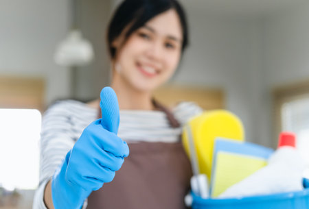 asian woman wearing apron holding cleaning products smiling happy and positive, thumb up doing excellent and approval signの写真素材