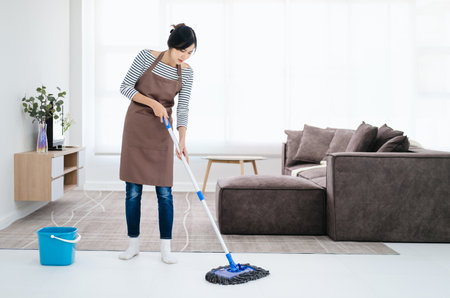 Pretty young lady in apron wiping floor with mop, doing house cleanup living room, free space. Positive housewife tidying her apartment, doing household chores. Professional sanitary service concept.の写真素材