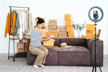 Vlogger, blogger small SME business asian beautiful woman using mobile phone video call for sell clothes live stream selling online, show product present detail at home office, entrepreneur ecommerceの写真素材