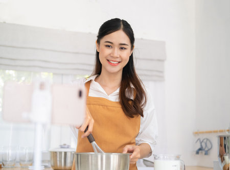 baker online courses. food preparation and culinary training class concept. smiling woman chef prepare ingredients in the kitchen and shooting video of herself using mobile phone on a tripod.の写真素材