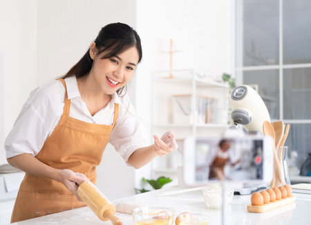 baker online courses. food preparing and culinary training class concept. smiling asian chef kneading dough in the kitchen and shooting video of herself using mobile phone on a tripod.の写真素材