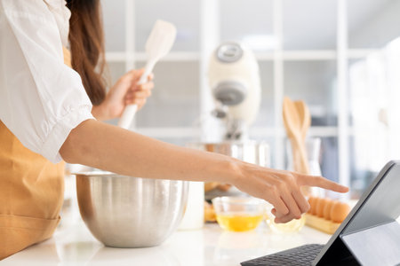 Close up of hands with computer in kitchen.Learn cooking online. A young woman is watching cooking tutorial video in the kitchen.Reading recipe while making cookies Following Recipe On Digital Tablet.の写真素材